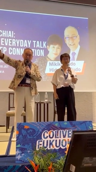 Hidehiko raising his arm with energy on stage beside Mikiko