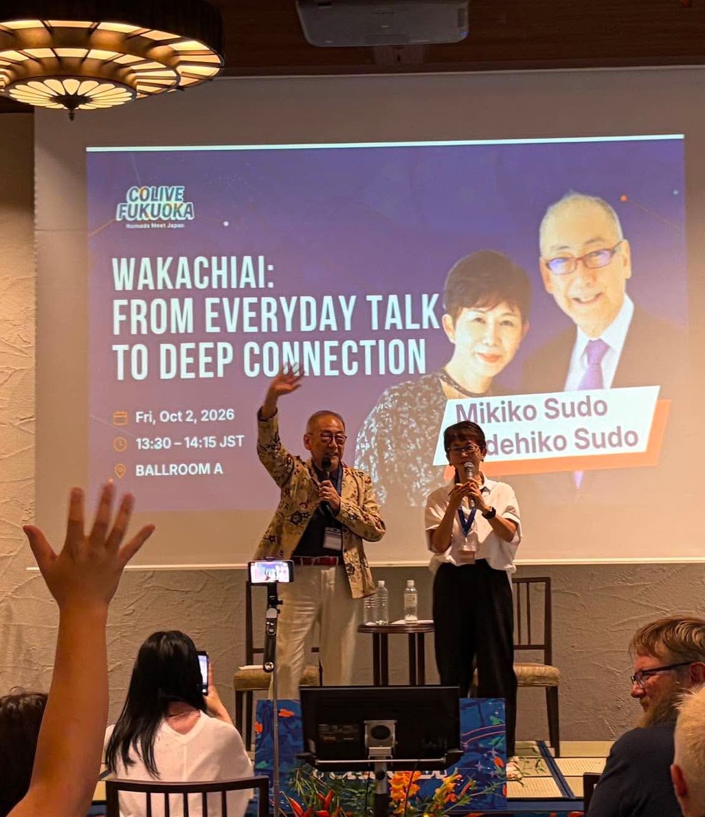 Hidehiko waving on stage beside Mikiko in front of the Wakachiai title slide, with audience hands raised at COLIVE Fukuoka